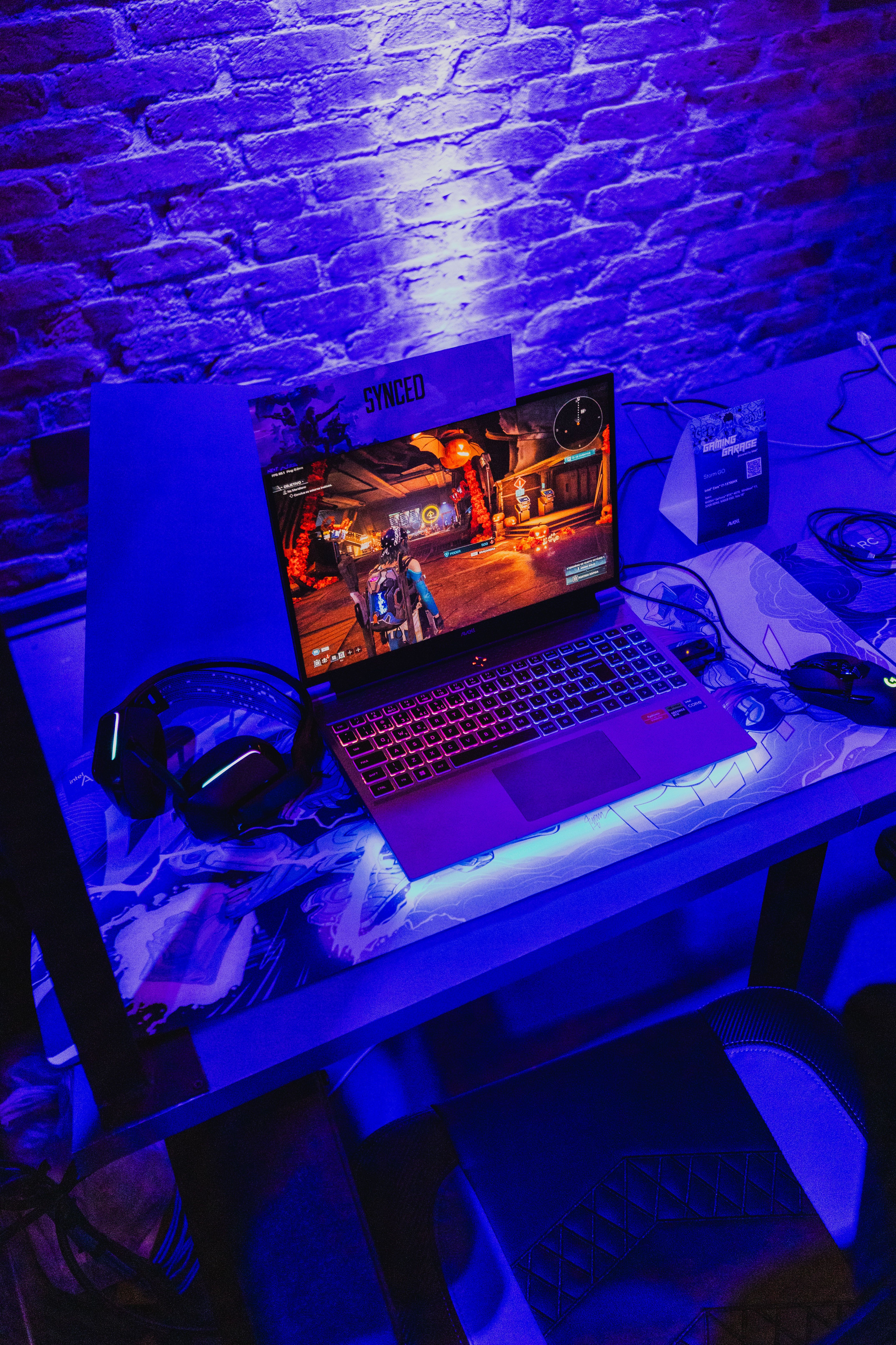 A gaming laptop showing a colorful game on a desk lit in blue, with a headset on the left and a gaming mouse on the right, in front of a brick wall