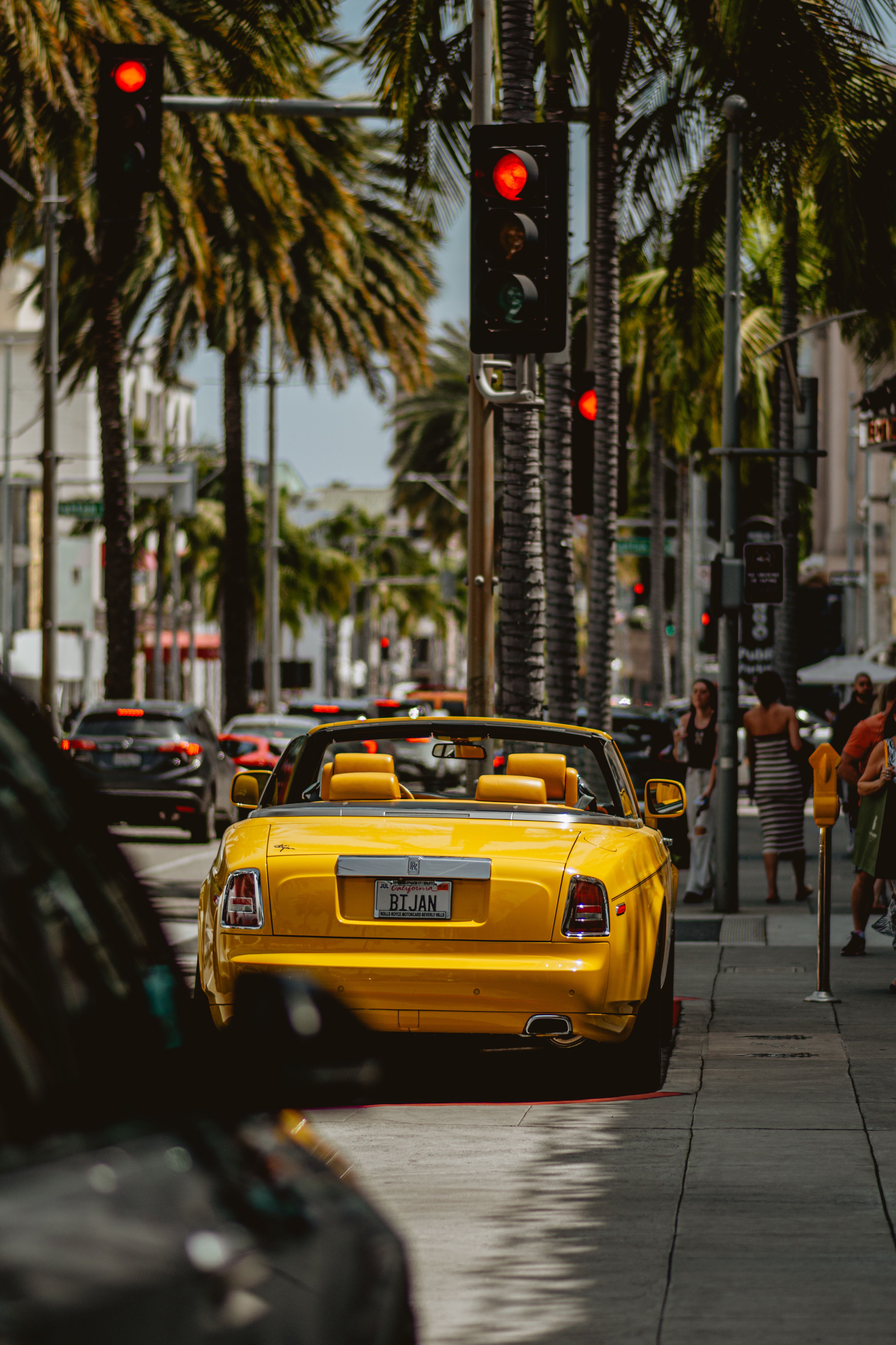 Yellow Rolls-Royce convertible with a tan interior parked on a palm-lined street at a red traffic light, seen from behind